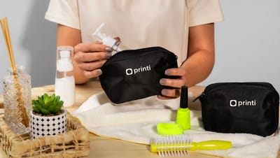 Necessaire personalizada preta com logo Printi sendo aberta para guardar frasco de sabonete e itens de cuidados pessoais.