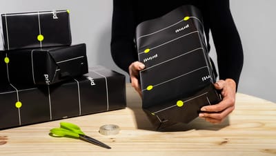 Pessoa embrulhando caixas de presente com papel de presente preto personalizado com detalhes gráficos em linhas brancas e pontos verde neon da Printi. As caixas aparecem sobre uma mesa de madeira junto com tesoura e fita adesiva