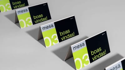 Display de mesa em formato prisma triangular personalizado, exibindo numeração de mesa “03” e mensagem “boas vindas!” em design moderno nas cores preto, branco e verde, apresentado em repetição sobre fundo neutro para destacar o material gráfico.