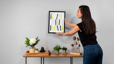 Quadro decorativo personalizado com moldura preta e arte gráfica moderna sendo colocado na parede por uma mulher.