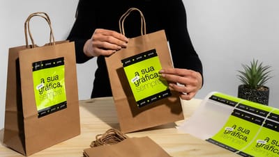 Aplicação de adesivo personalizado em sacola de papel kraft, com pessoa colando etiqueta verde da Printi na embalagem