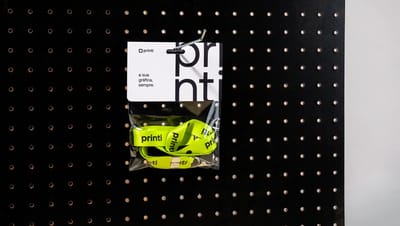 Solapa personalizada da Printi presa em painel perfurado exibindo cordão promocional verde-limão com logo Printi dentro de embalagem transparente, utilizada para apresentação de brindes, identificação de produtos e materiais promocionais em lojas e evento