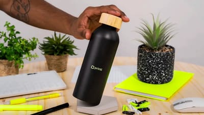 Squeeze térmico preto personalizado com tampa de bambu sendo segurado sobre mesa de escritório com caderno, canetas e plantas.