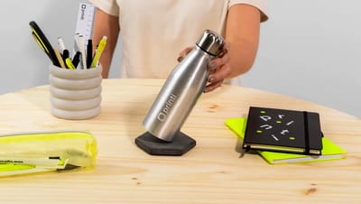 Squeeze metálico personalizado com logo Printi sendo segurado por uma pessoa sobre mesa de madeira, ao lado de caderno, estojo transparente com canetas e porta-lápis com materiais de escritório.